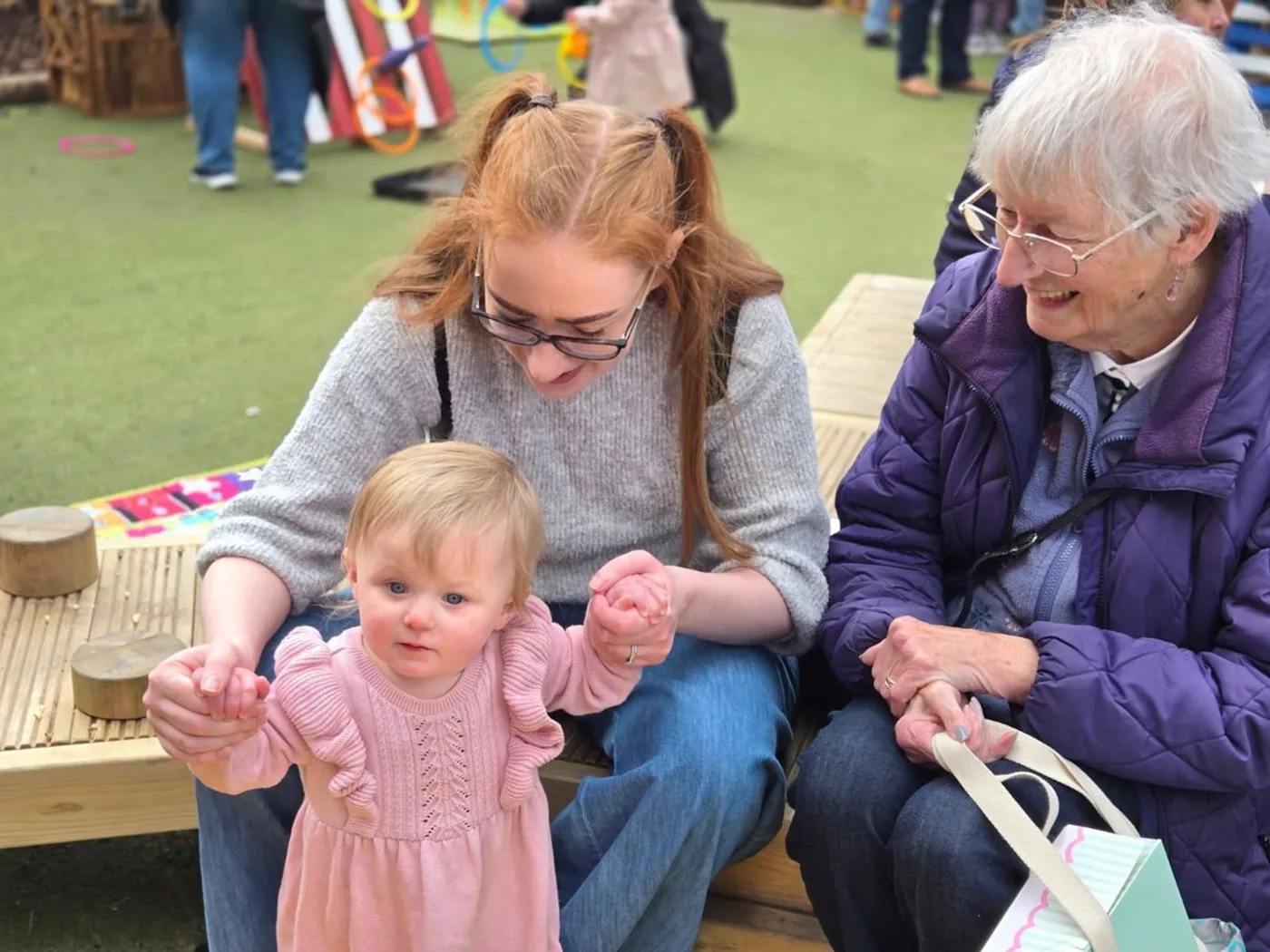 A woman holding a toddler's hands as she stands, with an older woman smiling beside them on a bench in the nursery garden
