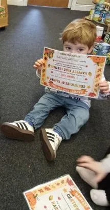 A young child sitting on the carpet holding up a Harvest Food Festival certificate of thanks
